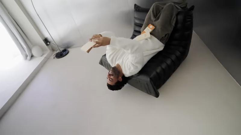 Le modèle Vincent Queijo allongé tête en bas dans un fauteuil, un emballage à la main, dans une pièce blanche